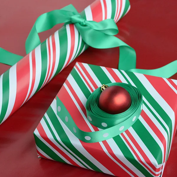 A red bauble surrounded by a green ribbon with white dots and sitting on top of a box wrapped in a red/green candy stripe design.