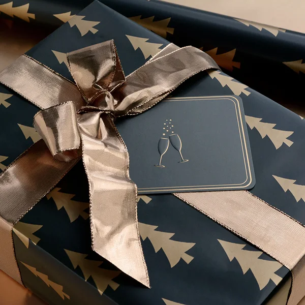 A box wrapped with a gold tree on black background wrapping paper, finished with a gold foil ribbon and a black/gold card with embossed champagne flutes.