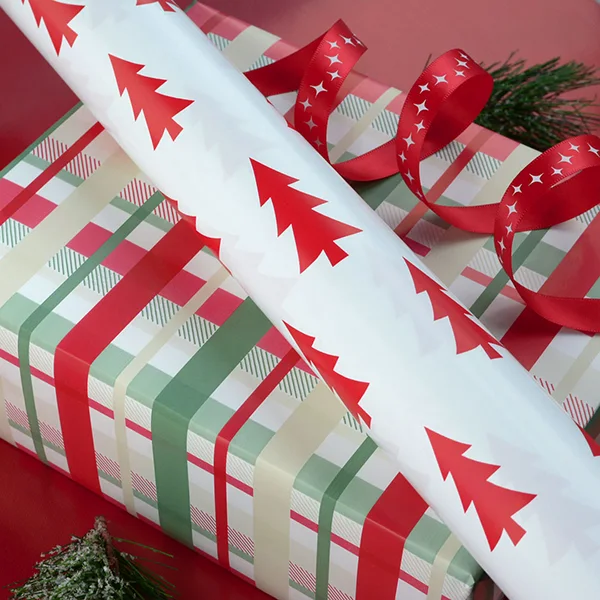 A roll of white wrapping paper with a red tree design on top of a wrapped box in check design. Also shown is a red satin ribbon with white stars.