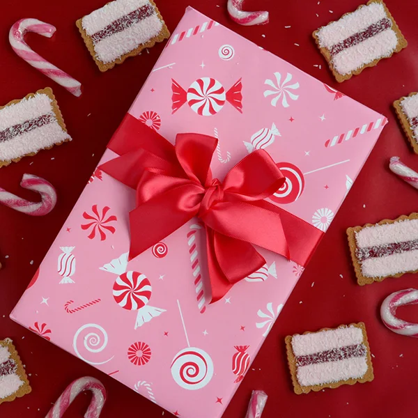 A box in a pink/red candy cane wrapping paper, finished with a red satin bow and surrounded by pink biscuits and candy cane.